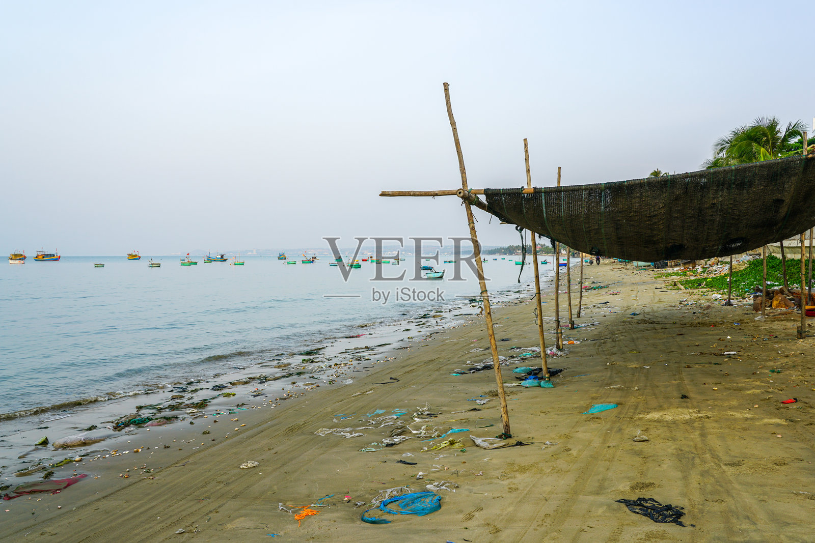 安静的钓鱼海滩上散落着垃圾，海上停着船只，还有简单的竹子结构提供阴凉。照片摄影图片