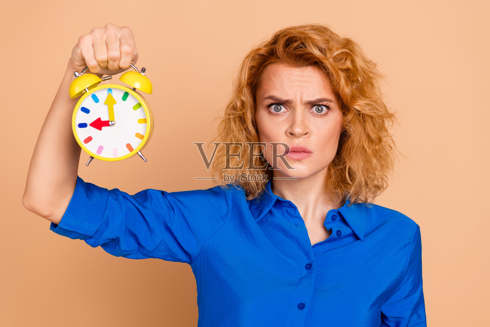 穿着蓝色衬衫的年轻女性手持黄色闹钟，面露紧迫感，背景为米色。照片摄影图片