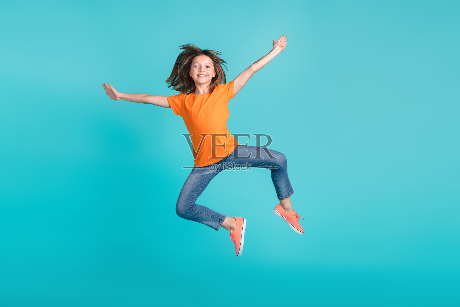 穿着时尚橙色衣服的快乐女孩在青色背景上开心地跳起，双手张开，享受拥抱和亲密的乐趣。照片摄影图片
