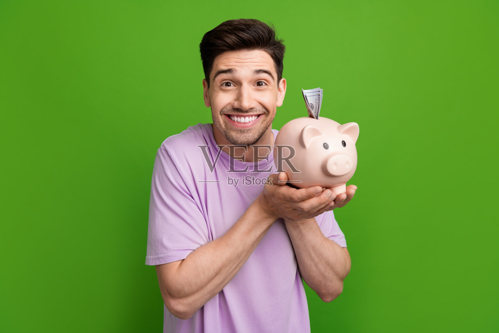 一位微笑着、露出牙齿的搞笑男子，棕色头发，穿着紫色T恤，手持储蓄罐靠近脸部，背景为绿色。照片摄影图片