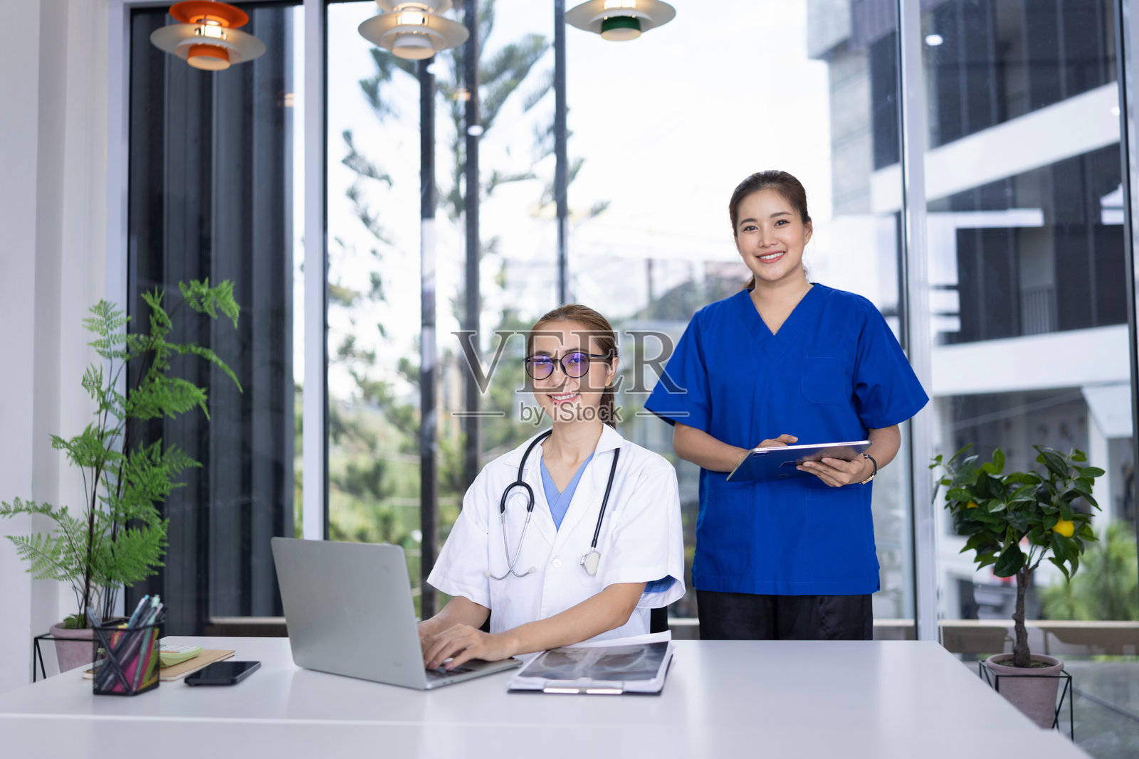 两位快乐的亚洲女性医生坐在医院的桌子前，手持笔记本电脑，面向镜头。照片摄影图片