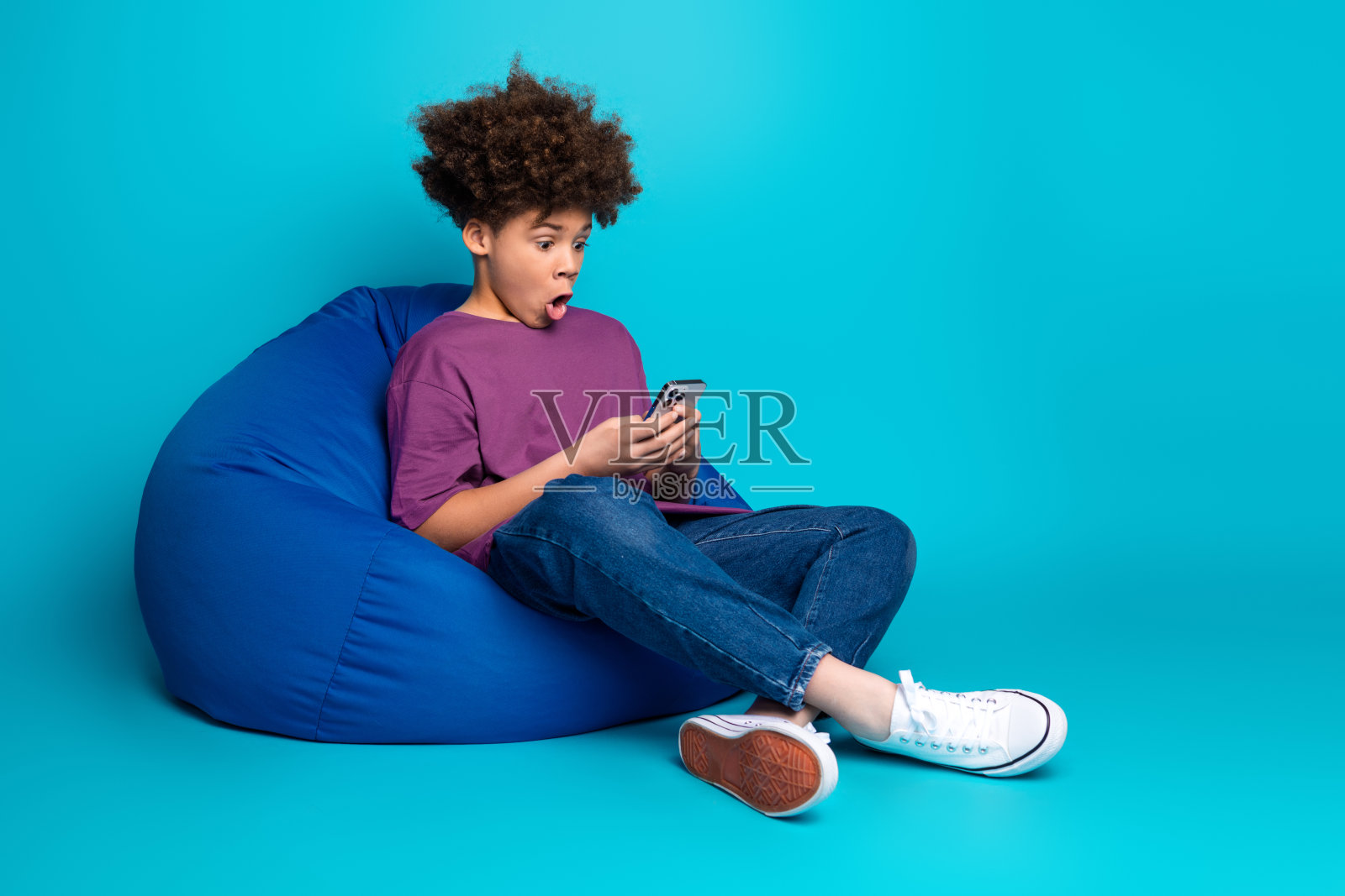 兴奋的青少年坐在豆袋椅上，手持智能手机，背景是充满活力的蓝色，展现现代生活方式。照片摄影图片