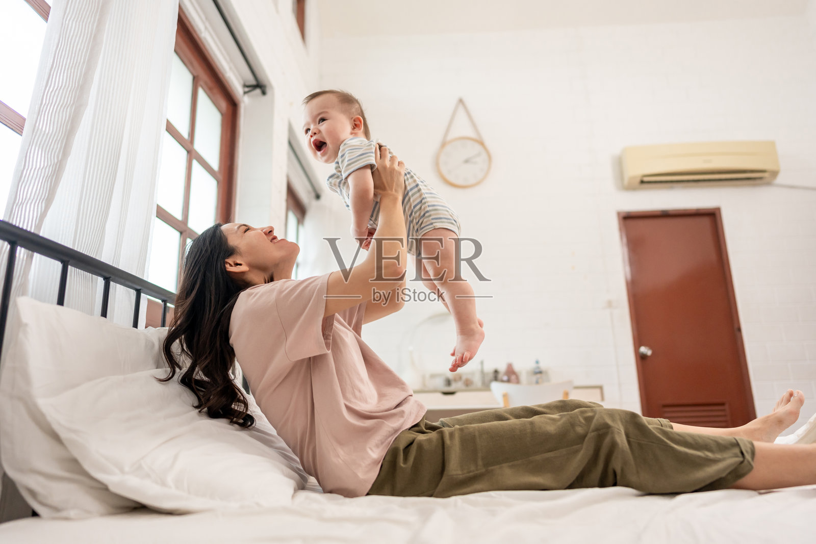 亚洲母亲在家中的卧室里轻轻地抱着并抛起她的婴儿男孩。迷人的美丽妈妈与新生儿在温馨的房间里分享温柔的时刻，营造出温暖而充满爱的家庭氛围。照片摄影图片