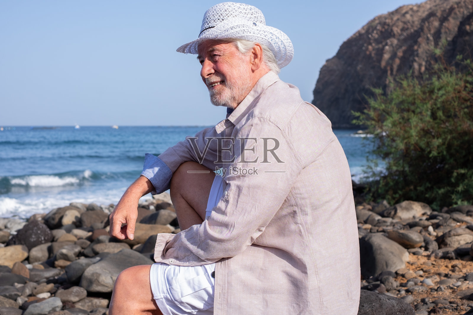 海边度假退休生活，放松的白胡子老人在岩石沙滩上坐着，目光远眺。戴着白色帽子的老爷爷在户外享受闲暇时光。照片摄影图片