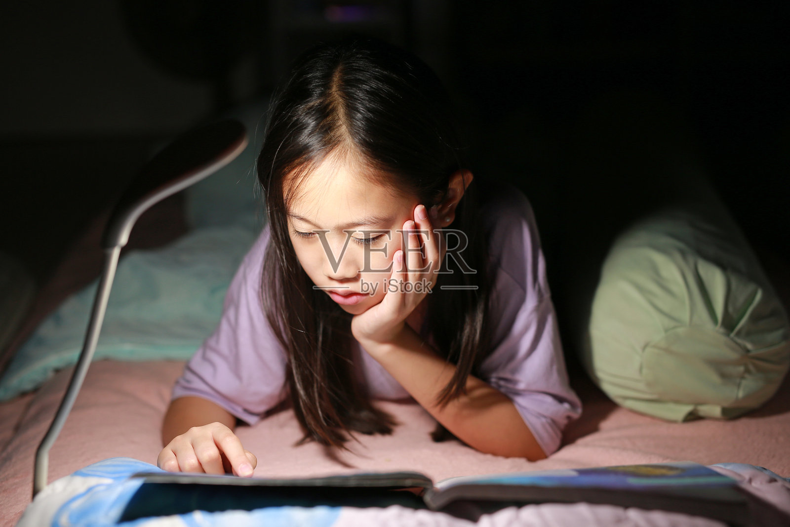 夜晚，亚洲女孩在床上躺着，灯光下专心阅读一本书。柔和的焦点。照片摄影图片