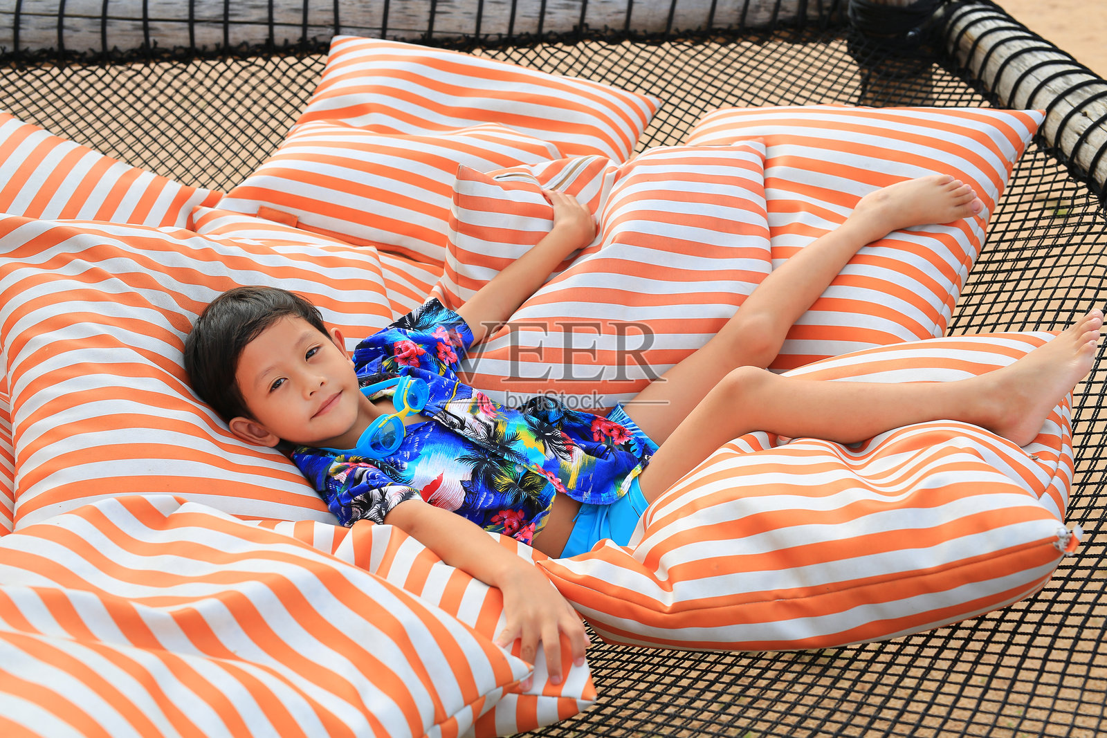 微笑的亚洲男孩穿着夏季服装，靠在大网状吊床的枕头上，吊床由耐用的绳索或线制成，悬挂在海滩上。照片摄影图片