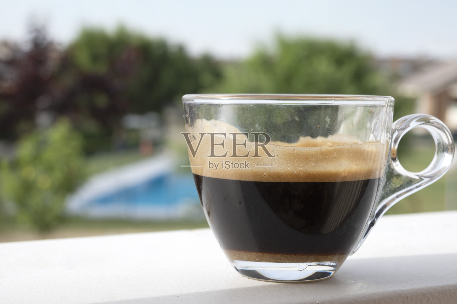白色表面上的一杯浓缩咖啡，背景是花园和游泳池。照片摄影图片