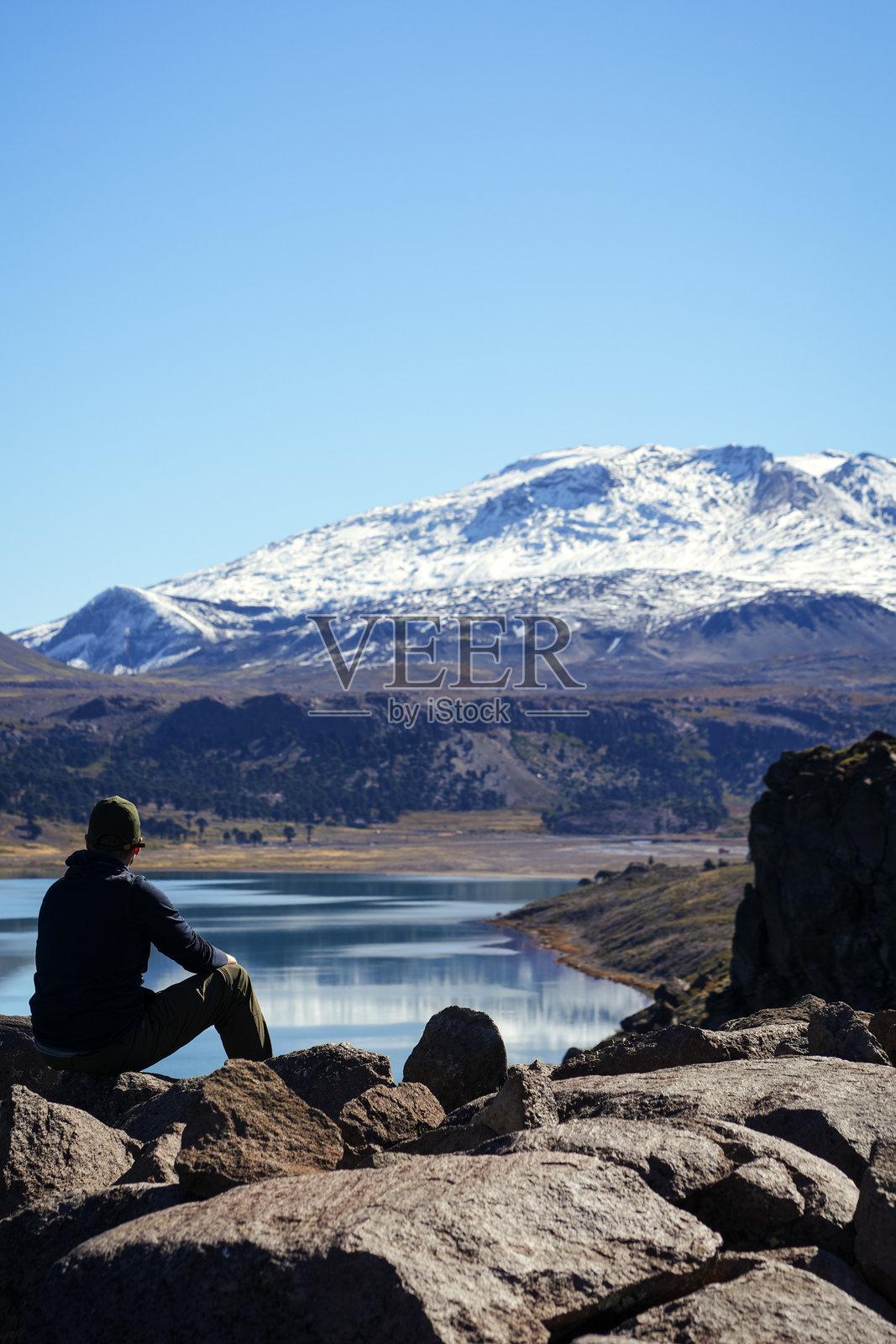 一个无法辨认的男人坐在岩石上，望着卡维亚湖和背景中的雪山科帕乌火山。阿根廷，内乌肯。照片摄影图片