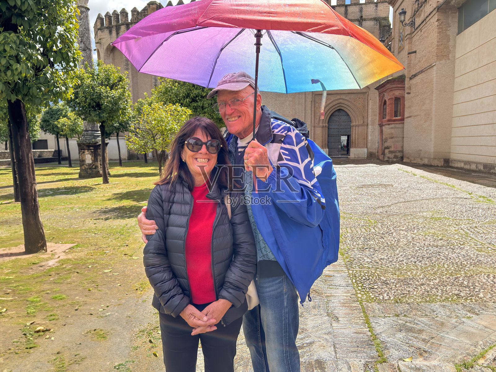 在塞维利亚的一座历史悠久的房子花园里，一对微笑的老年夫妇在伞下享受观光旅行的文化和愉快的退休生活。照片摄影图片