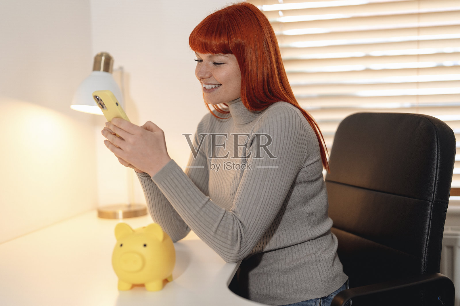 年轻女子微笑着使用智能手机，旁边是一个储蓄罐，坐在温馨的室内工作空间中，早晨的自然光洒入。照片摄影图片