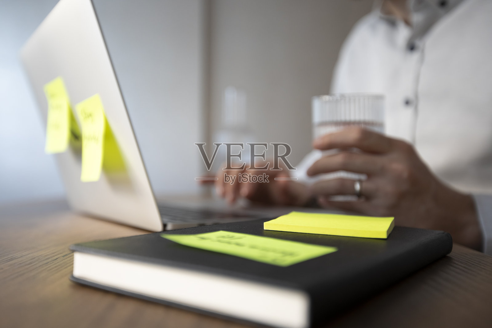 一个戴眼镜、穿白衬衫的男人微笑着，手里拿着一杯水。他的笔记本电脑上有两个弹出便签：“记录会议笔记”和“保持水分 :)”。桌子上有一个白色花盆里的小仙人掌。照片摄影图片