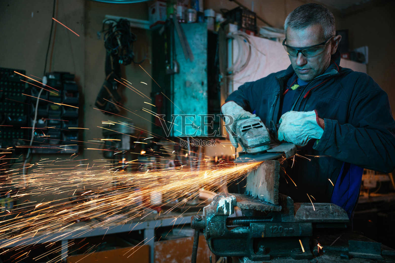 在昏暗的车间里，工业工人佩戴着防护眼镜，操作着角磨机，周围飞溅着火花。照片摄影图片