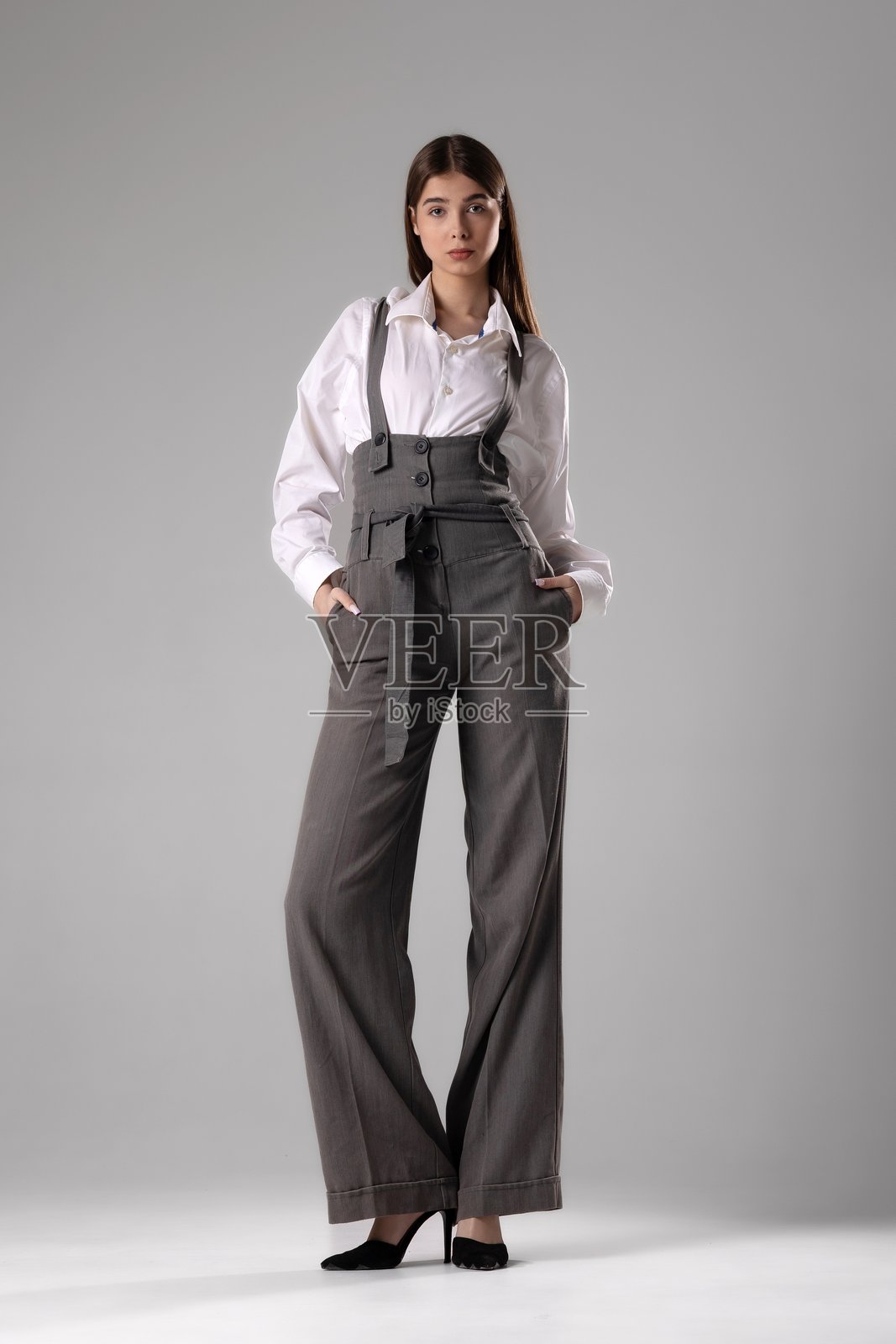 优雅的模特自信地穿着时尚的灰色工装和白衬衫，背景为简约的灰色。照片摄影图片