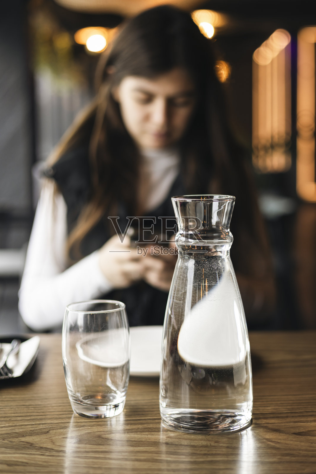 一壶水和一杯子，背景是一位在餐厅的女性。照片摄影图片