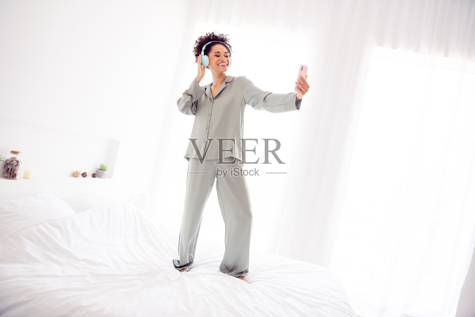 积极的博主女士在卧室床上用耳机拍摄早晨视频，穿着丝滑的睡袍，室内光线明亮。照片摄影图片