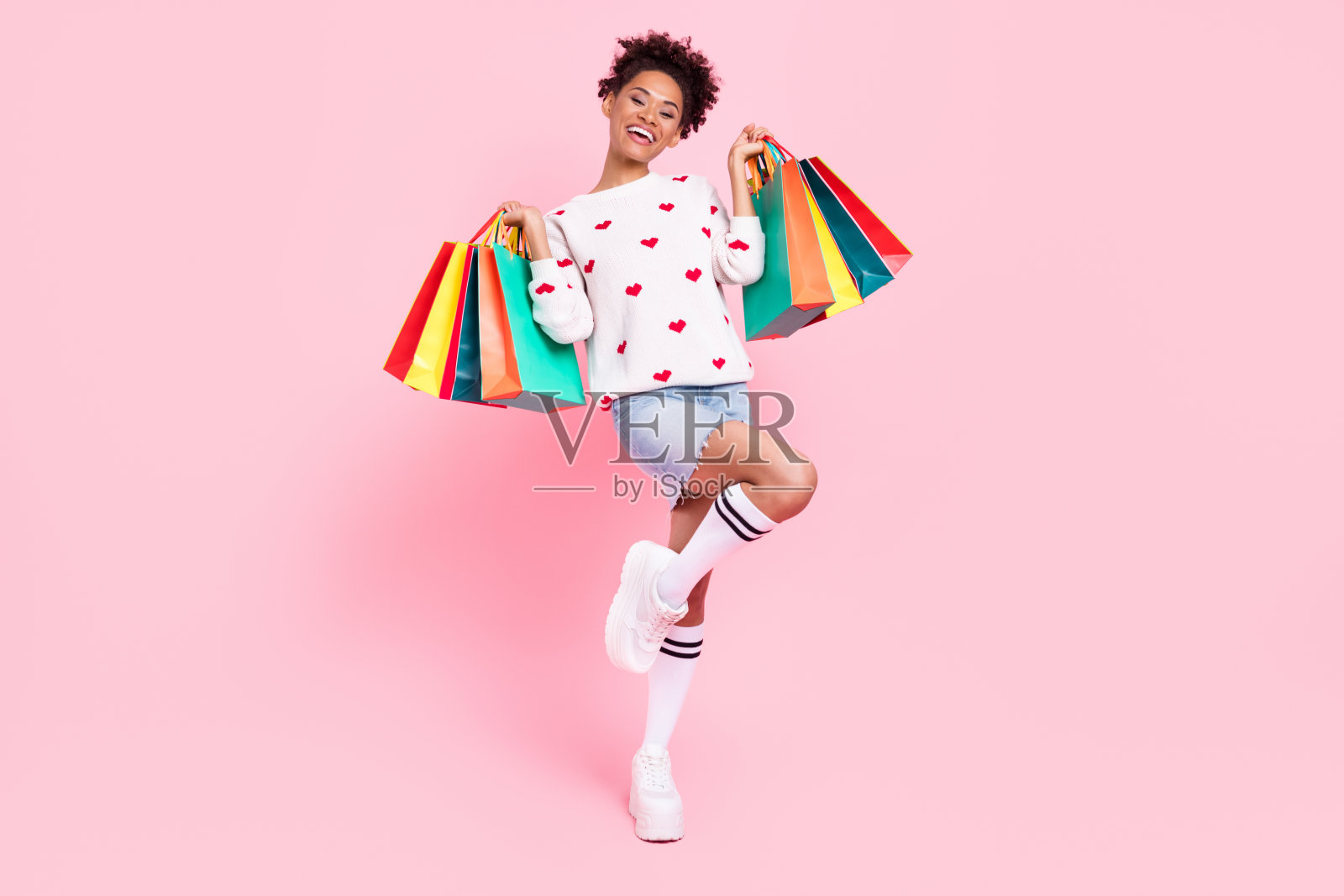 时尚的棕发年轻女士全身照，穿着毛衣、迷你裙、袜子和运动鞋，背景为粉色。照片摄影图片