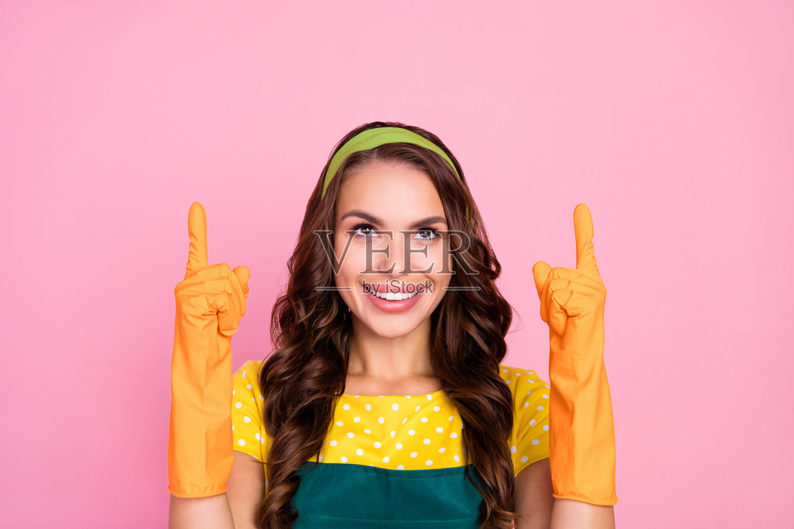 穿着绿色围裙和橡胶手套的甜美迷人的年轻女士，微笑着指向空旷的地方，背景为粉色。照片摄影图片