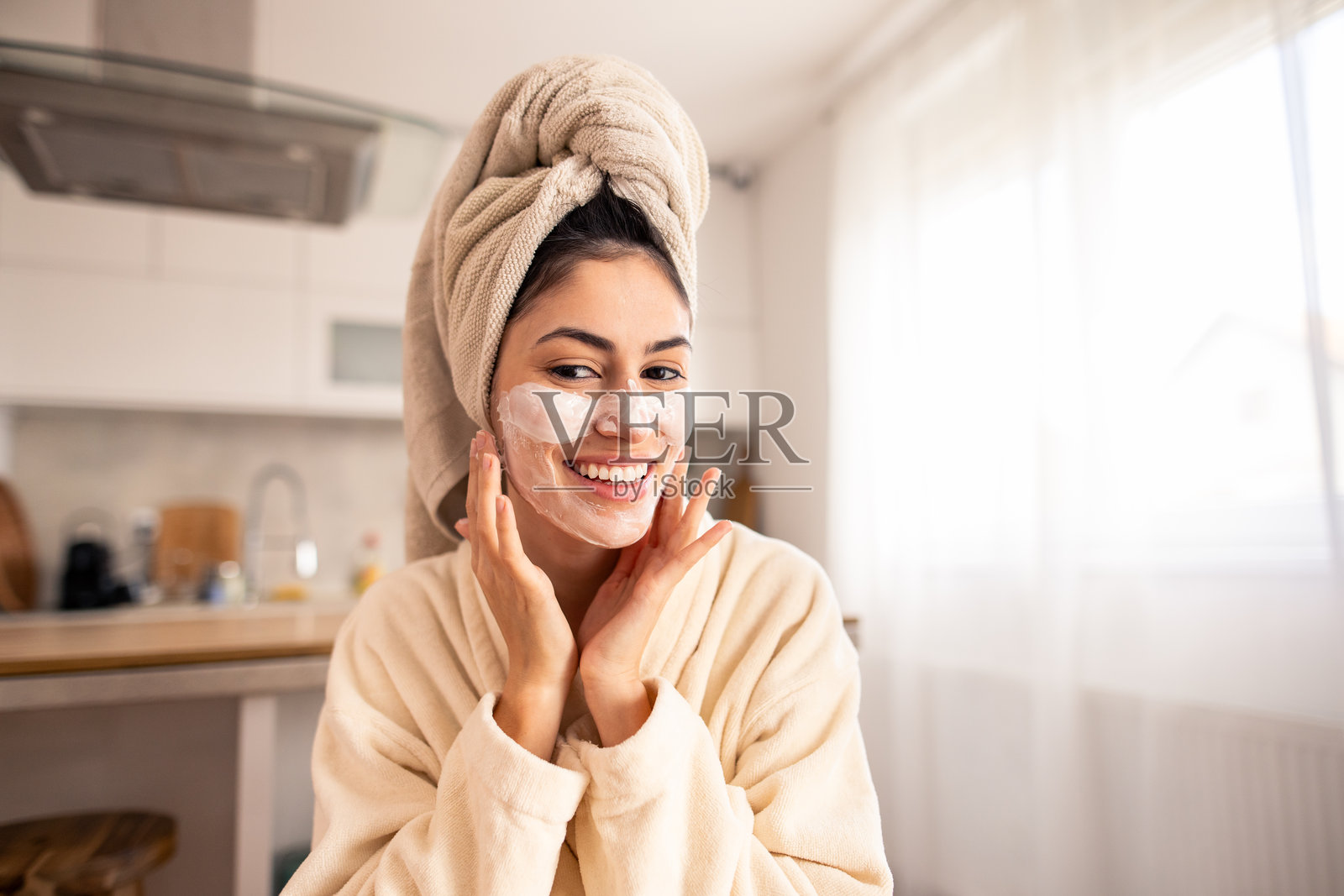 微笑的女性穿着米色浴袍和毛巾，在阳光明媚的厨房里涂抹护肤品照片摄影图片