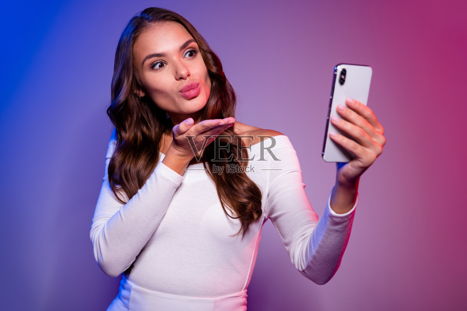 浪漫的女士拿着手机自拍，送出空气之吻，穿着衬衫，背景是霓虹渐变色。照片摄影图片