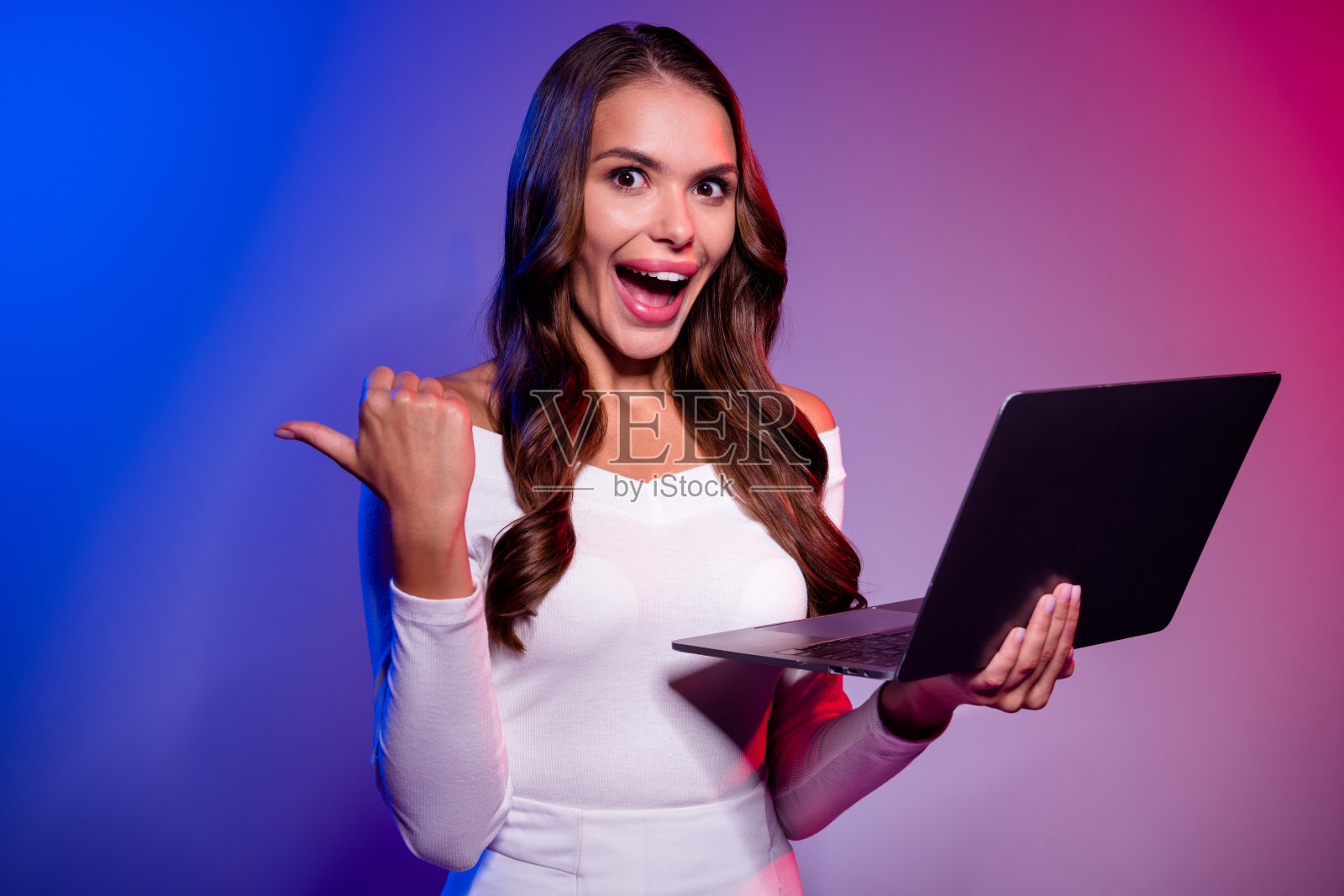 女士穿着白衬衫，手持电脑，指向在线游戏比赛横幅，背景为霓虹渐变色。照片摄影图片