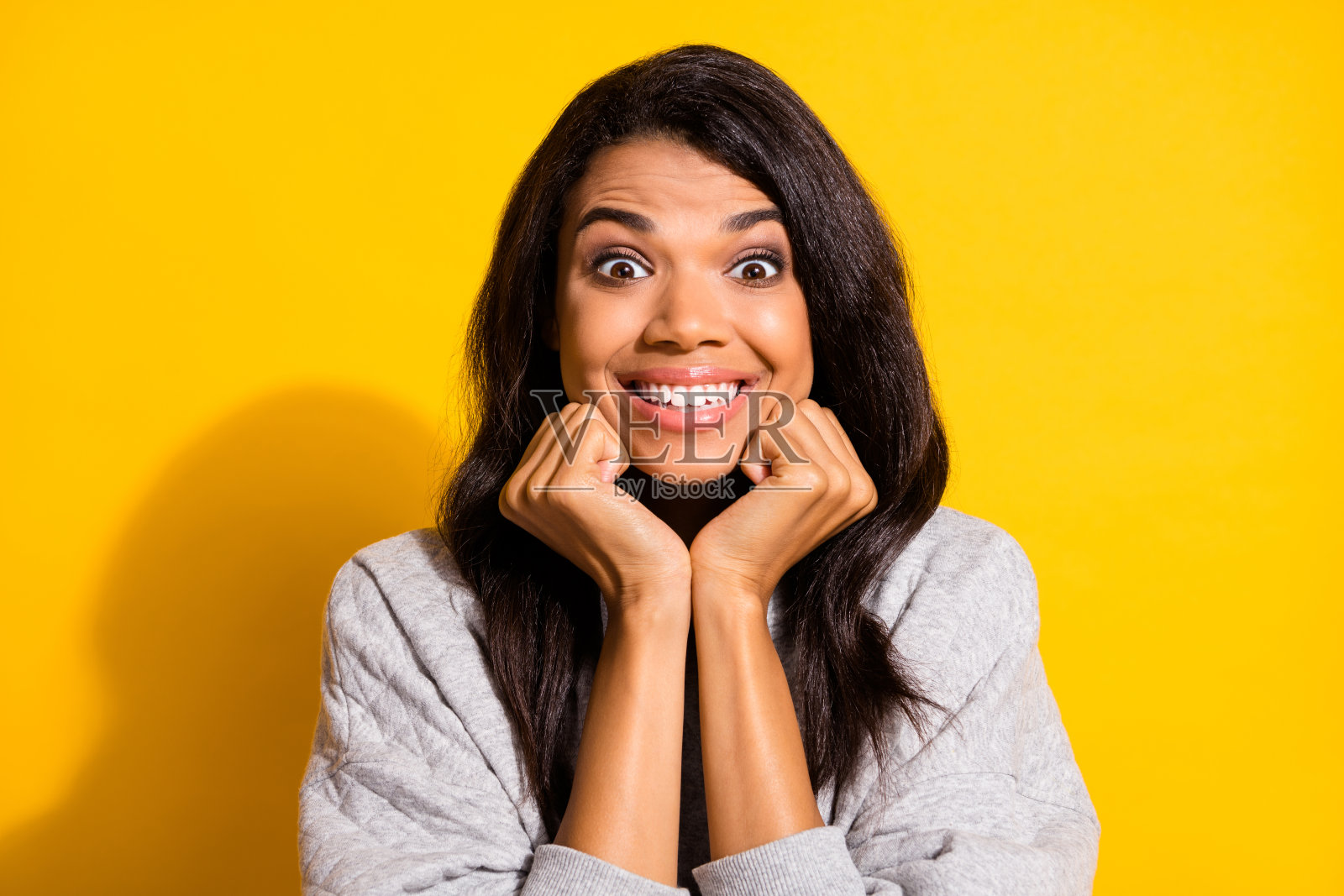 迷人的兴奋的深肤色女性穿着灰色连帽衫，双臂托着下巴，背景为黄色。照片摄影图片