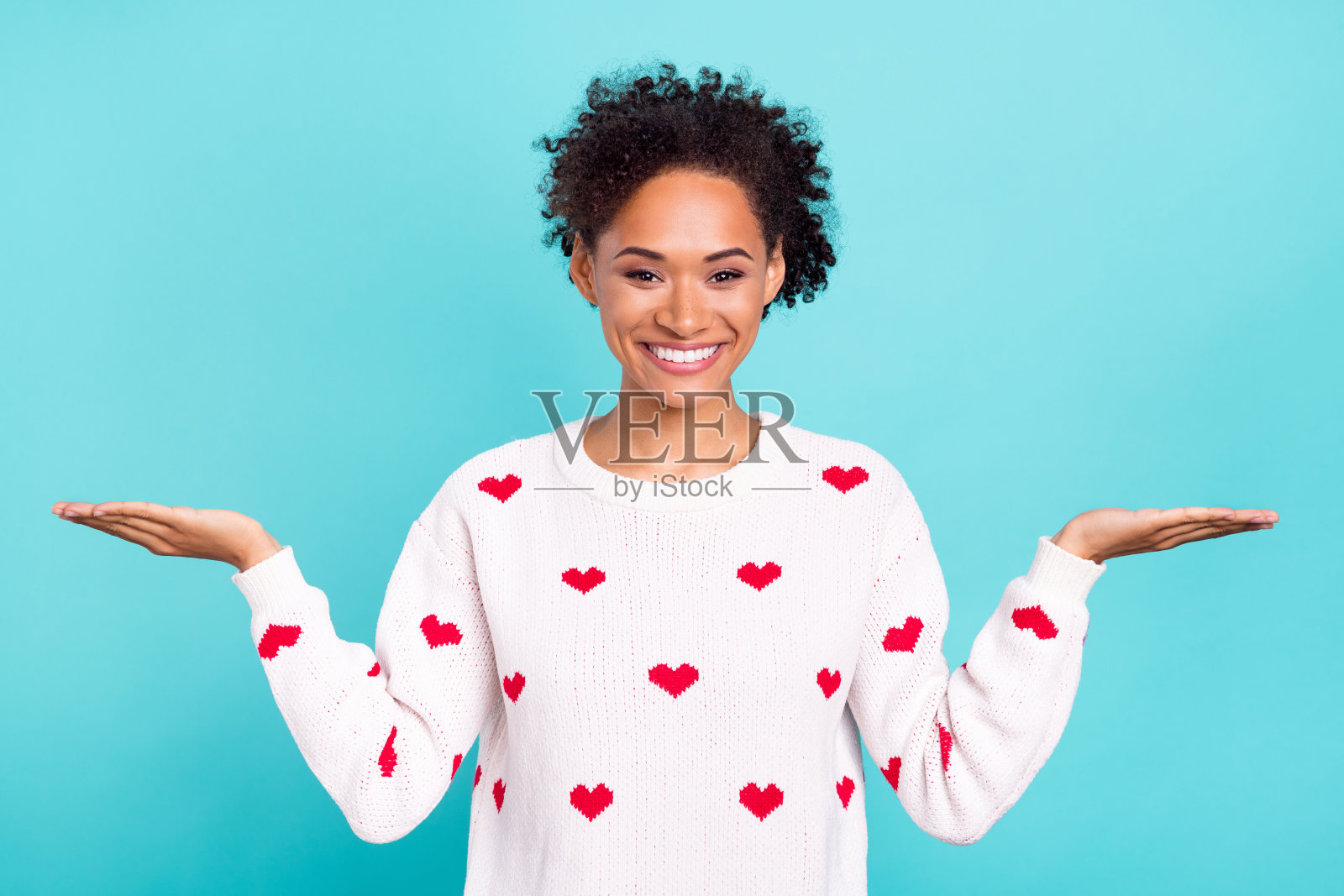 可爱的迷人非洲女孩穿着心形图案的毛衣，微笑着展示手臂，背景为空白的蓝色。照片摄影图片