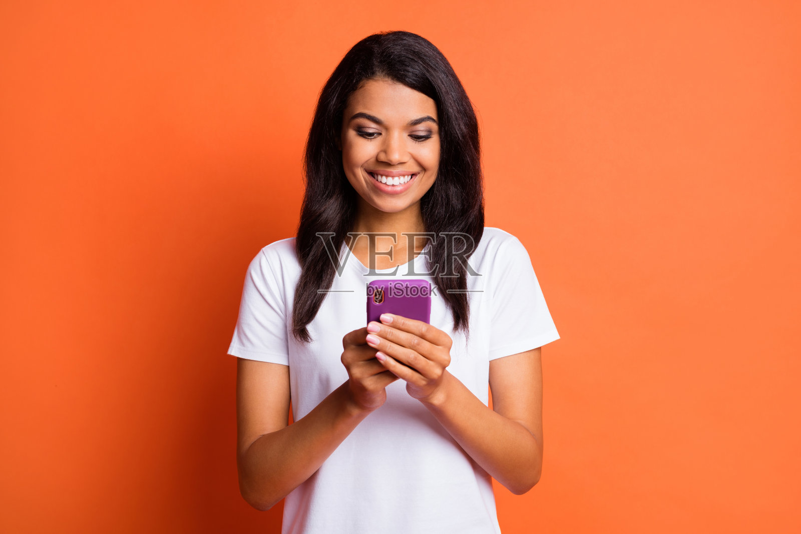 乐观的女孩穿着白色T恤，手持电话，背景为橙色。照片摄影图片