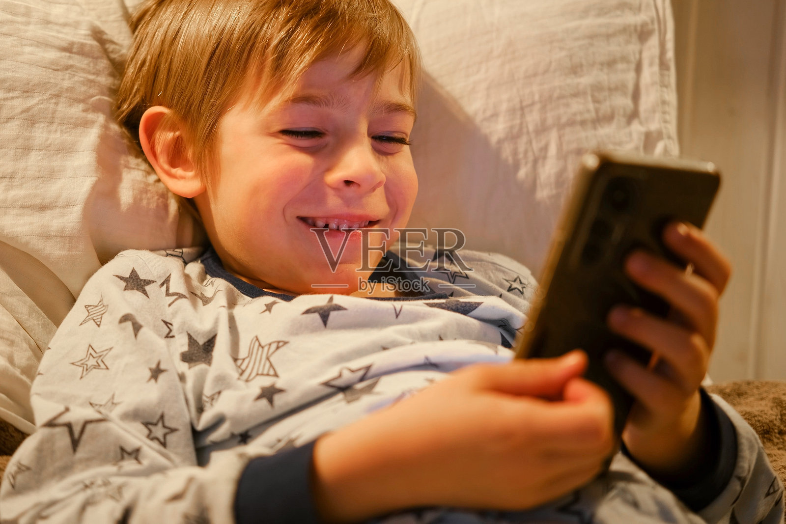 可爱的小男孩穿着睡衣躺在床上看智能手机，面带微笑。儿童的屏幕时间。孩子在晚上玩手机游戏。社交媒体成瘾。童年，现代科技。照片摄影图片