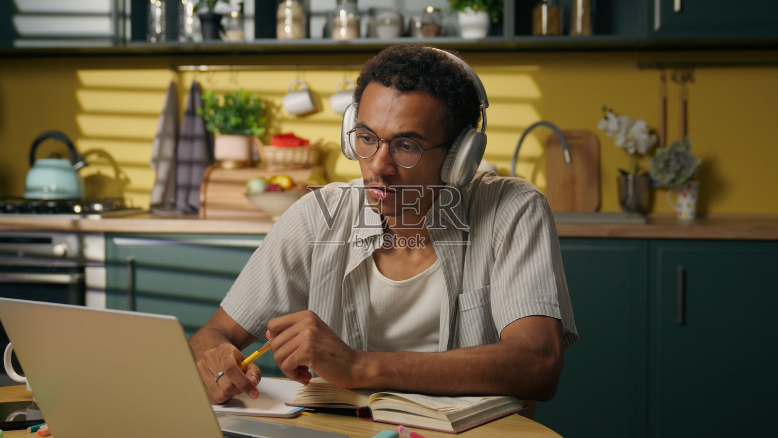 非裔美国商人男士，戴着耳机，正在进行视频通话会议，使用笔记本电脑，男性学生在家厨房远程学习，在线上课，老师在写作业笔记。照片摄影图片