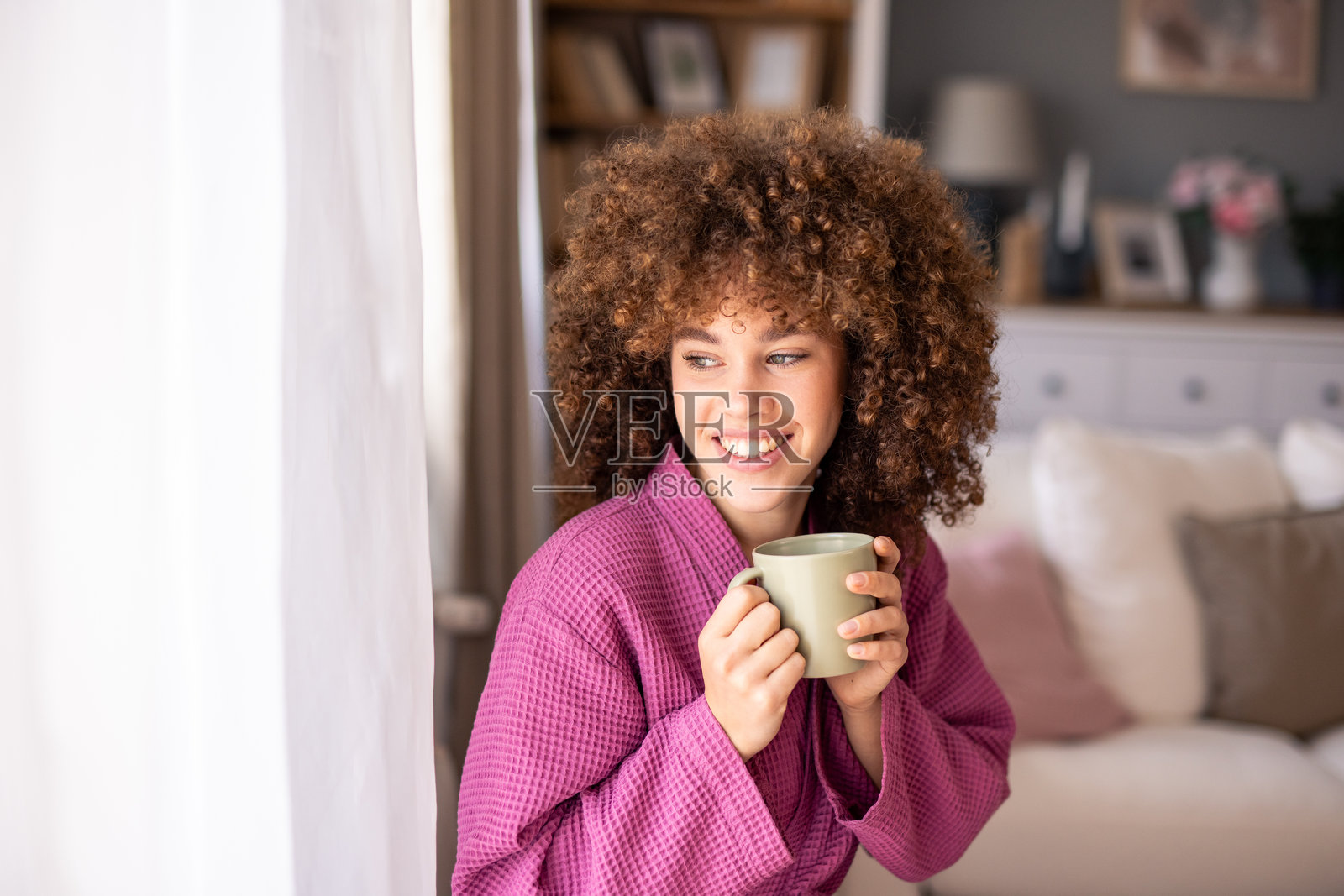 愉快的白人女性穿着粉色浴袍，在室内放松，手中捧着一杯温暖的茶。照片摄影图片
