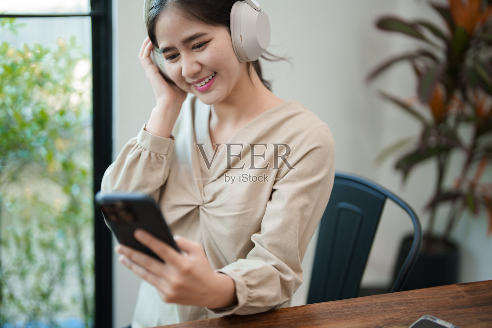 一位年轻女性坐在桌子前，戴着耳机，微笑着看着手机。她似乎在享受一段对话或音乐，工作在一个现代而明亮的办公室环境中。照片摄影图片