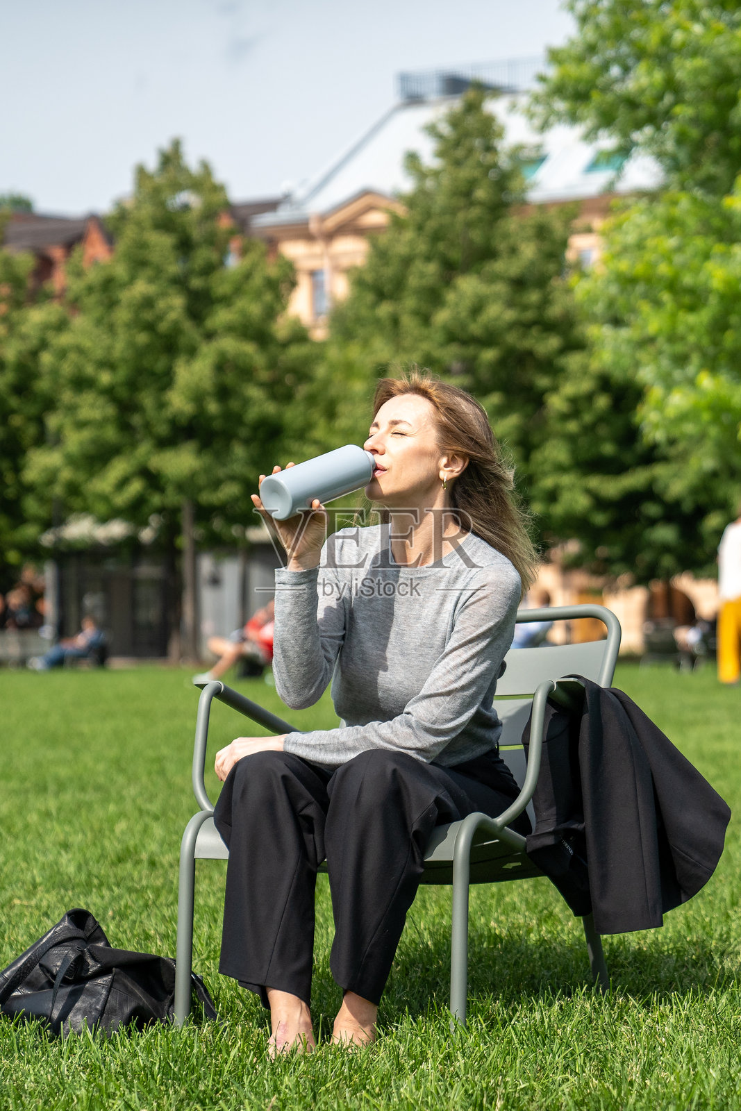 疲惫的女人在公园的椅子上休息，微微眯着眼睛喝水，坐在草坪上。照片摄影图片