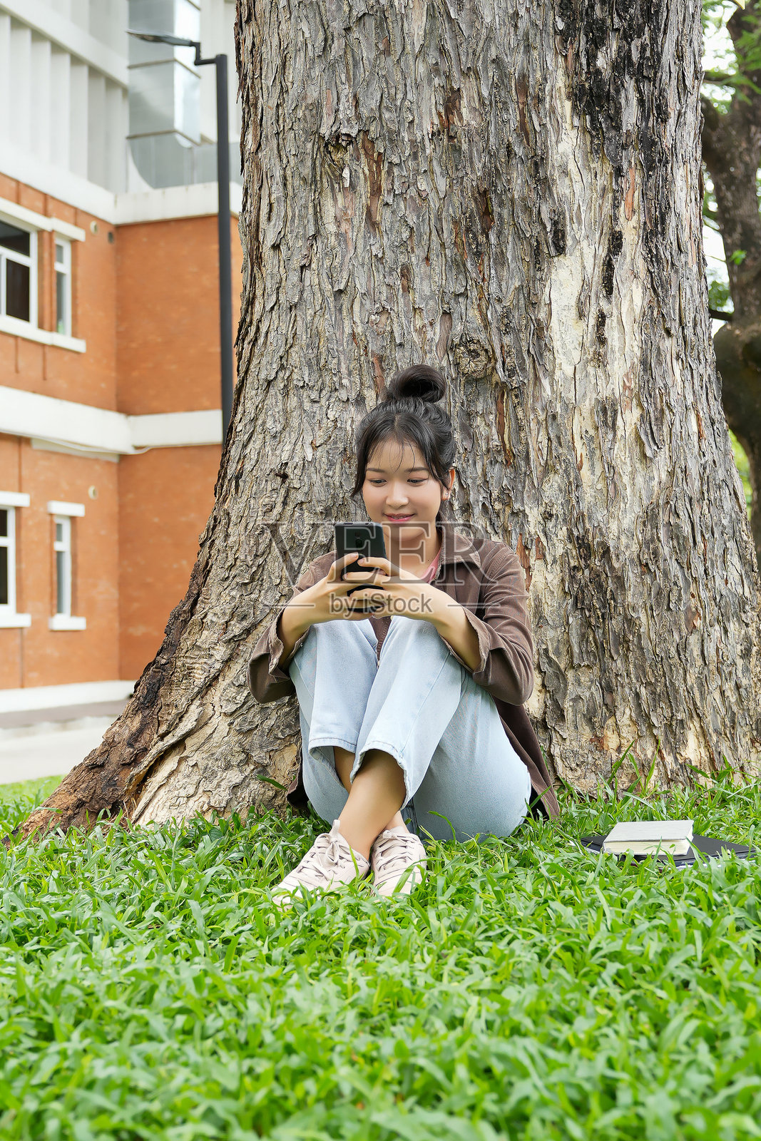 一名高中生坐在树下的草地上，微笑着看着她的智能手机。她穿着棕色夹克和浅蓝色牛仔裤，看起来放松而快乐。照片摄影图片