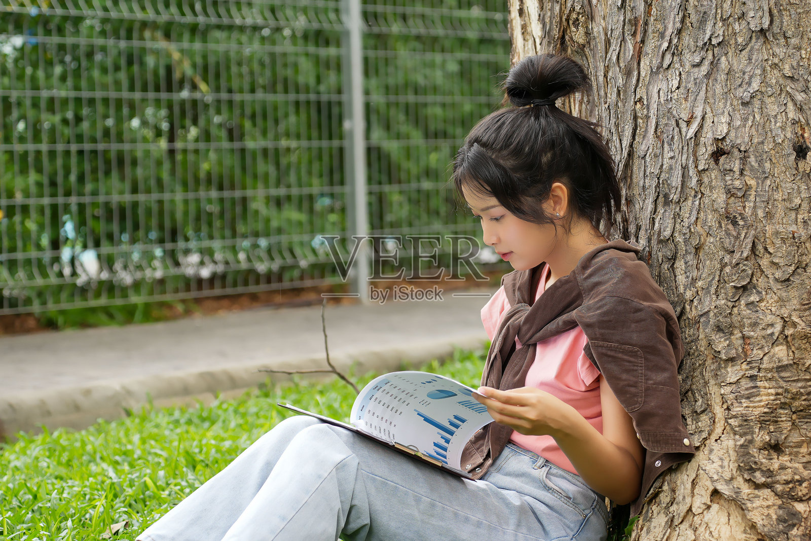 一名高中生靠在树上，专心致志地学习。她专注于在笔记本上写字，同时手里拿着一本书。照片摄影图片