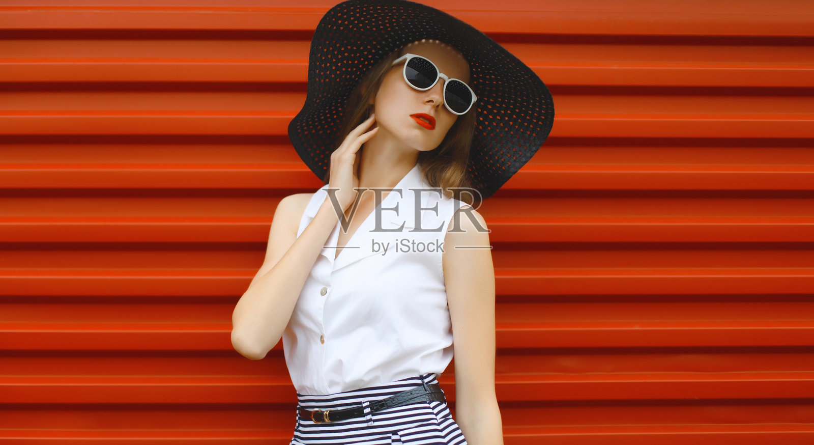 身穿夏季黑色帽子的美丽时尚年轻女性，时尚女孩模特站在红色背景前。照片摄影图片