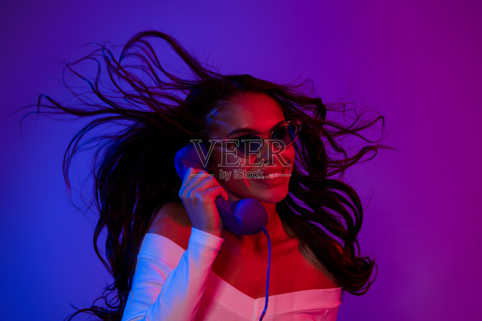 性感的年轻棕发女士穿着白色连衣裙，戴着眼镜，独自在霓虹色背景下打电话。照片摄影图片