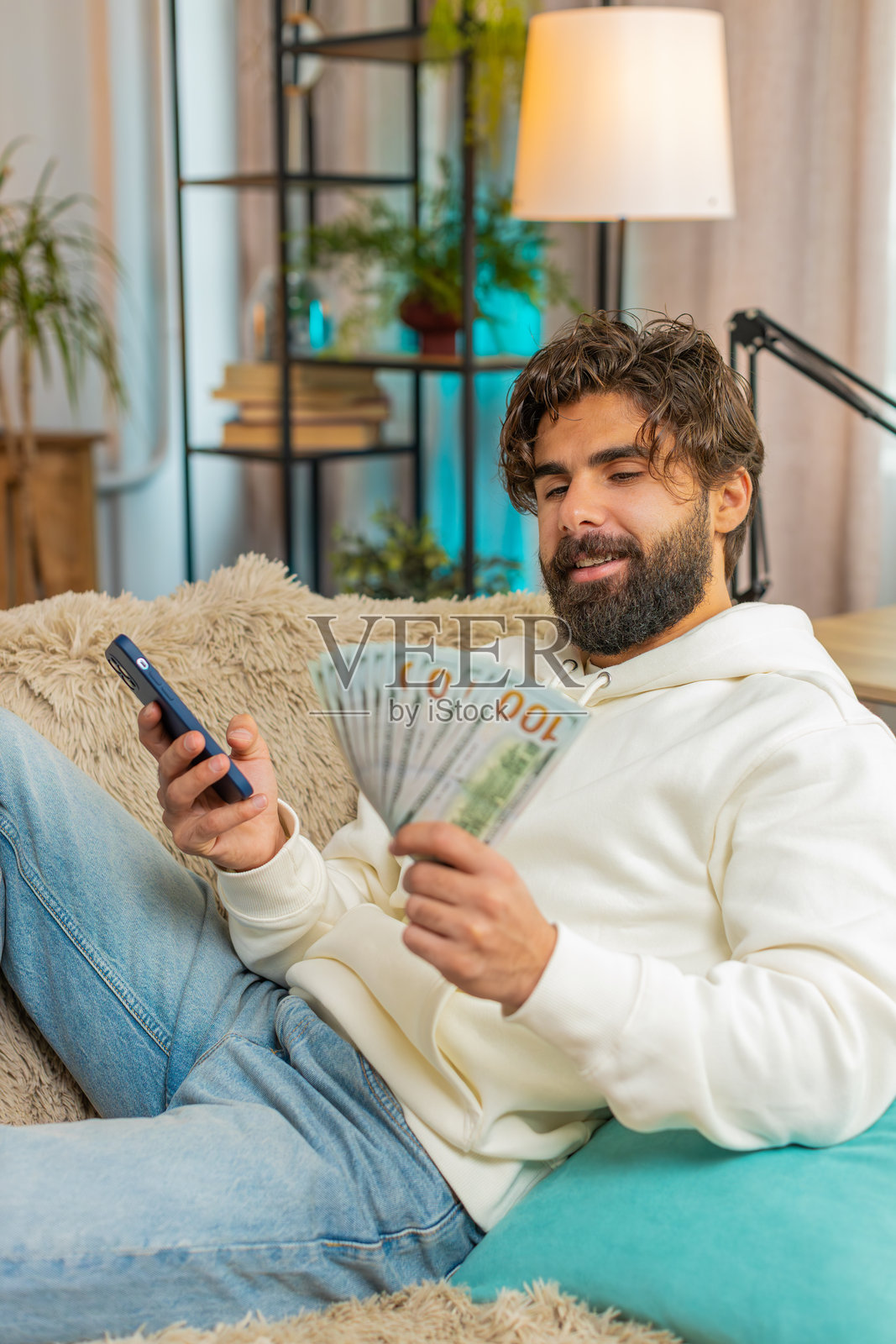 成功的富有年轻印度男子坐在家里的沙发上，手里拿着美元钞票和智能手机。照片摄影图片