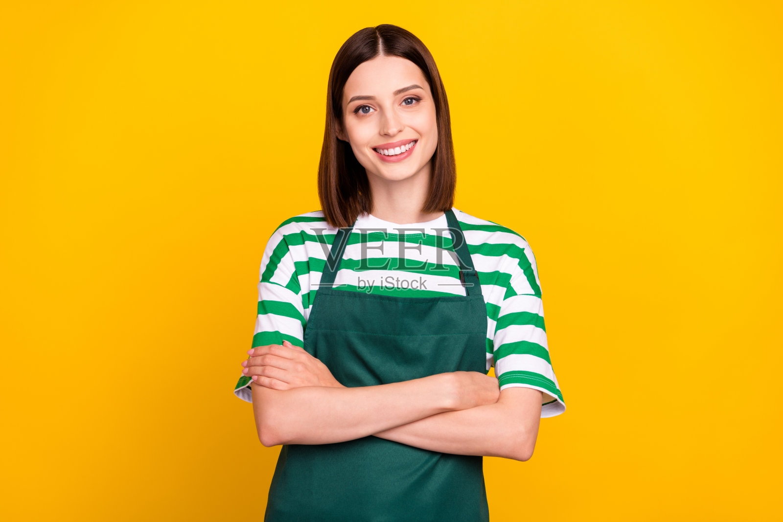 可爱的棕发年轻女造型师双臂交叉，穿着围裙和条纹T恤，背景为黄色。照片摄影图片