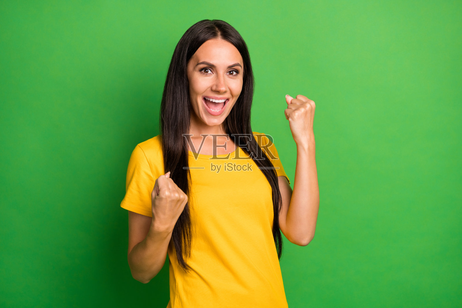 迷人快乐的年轻女性举起拳头庆祝胜利，穿着黄色T恤，背景为明亮的绿色。照片摄影图片