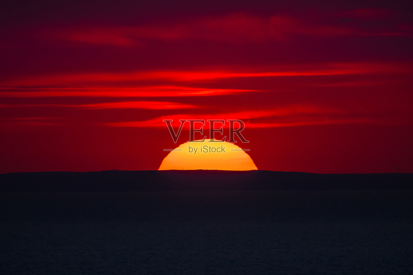 海平线上绚丽的红色日落，太阳半掩于远处山丘的黑色轮廓后。照片摄影图片