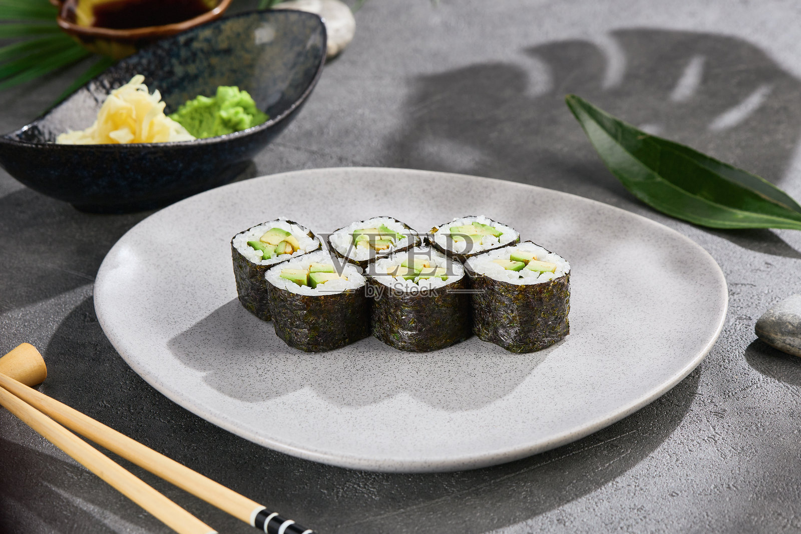极简主义的素食鳄梨细卷，放在灰色陶瓷盘上，配有筷子和芥末。现代日本食品造型，利用自然光和阴影效果，适合健康菜单或餐厅横幅概念。照片摄影图片