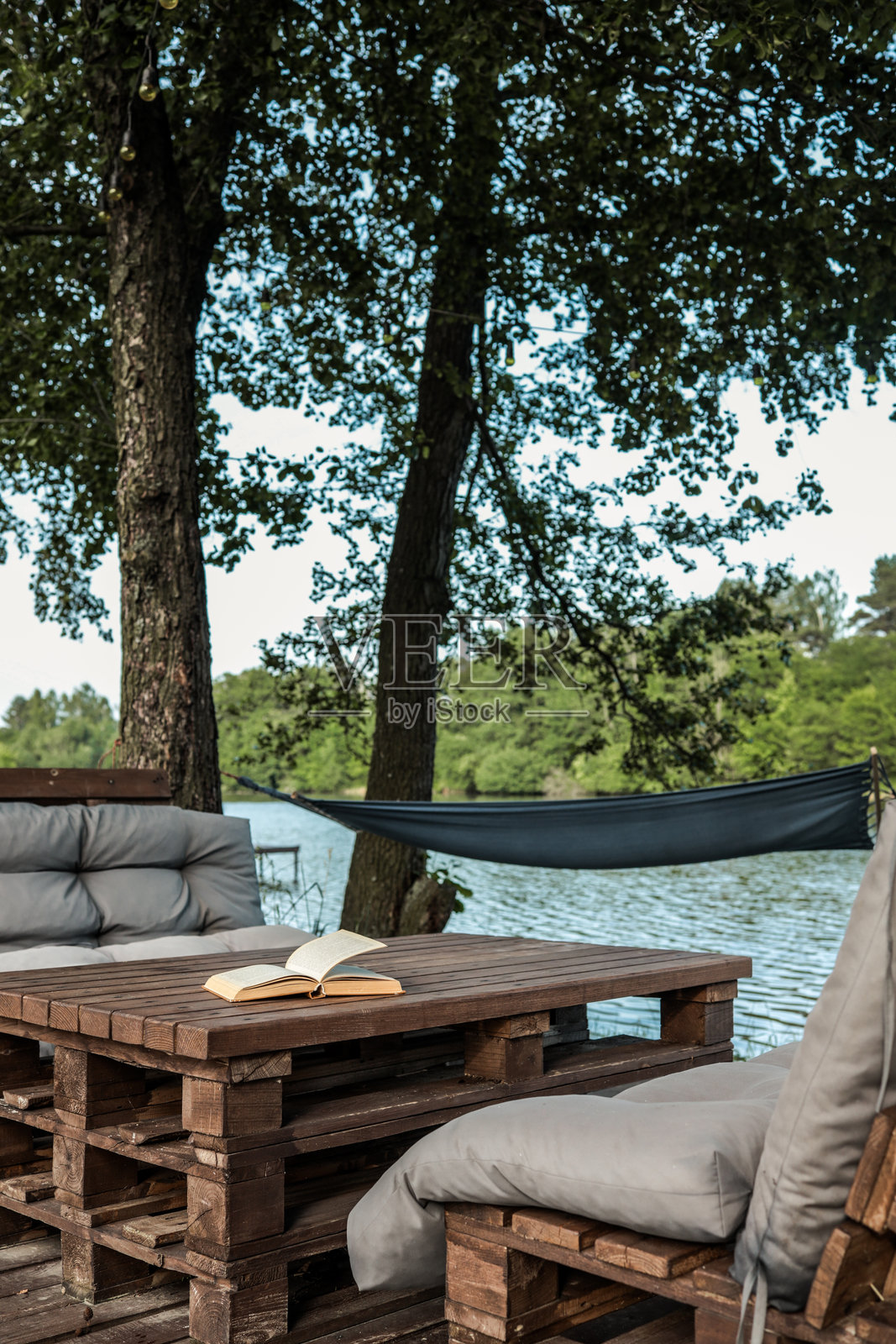 在宁静的湖边，古老的书籍摊开在一张质朴的木桌上，背景中有吊床，四周环绕着绿意盎然和舒适的户外垫子。照片摄影图片