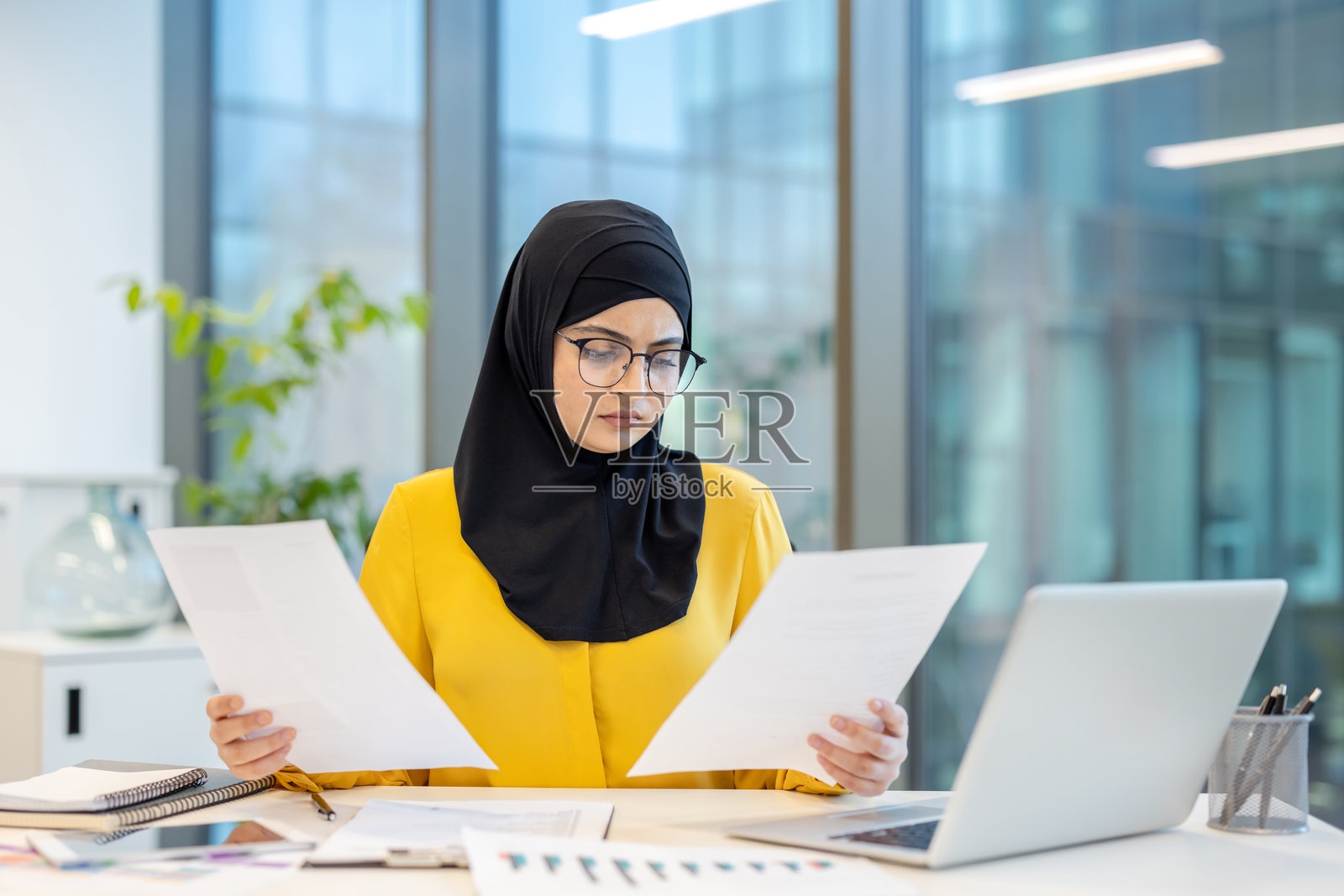 一位阿拉伯女商人在现代办公室环境中审阅文件，佩戴着头巾和眼镜。照片摄影图片