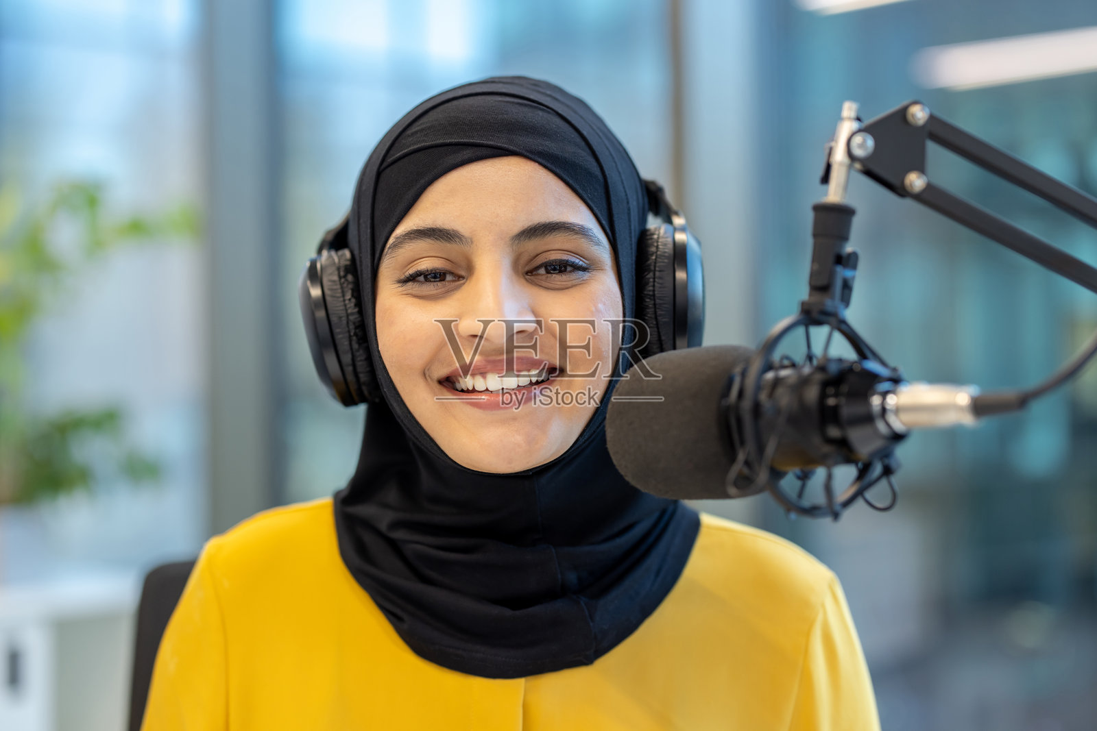 一位微笑的穆斯林女性戴着头巾在现代录音室录制播客。照片摄影图片