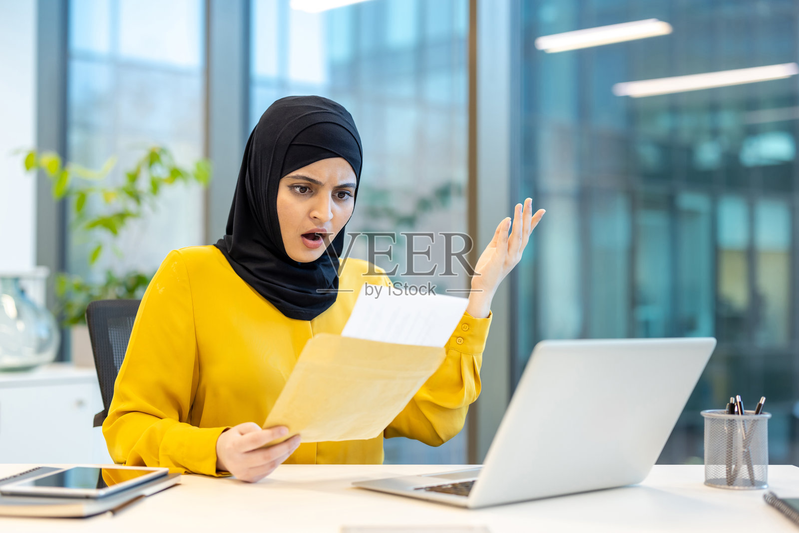 戴着头巾的女性收到坏消息的电子邮件通知。阿拉伯办公室工作人员感到沮丧和失望，手中拿着纸质信封，身处办公室工作环境中。照片摄影图片