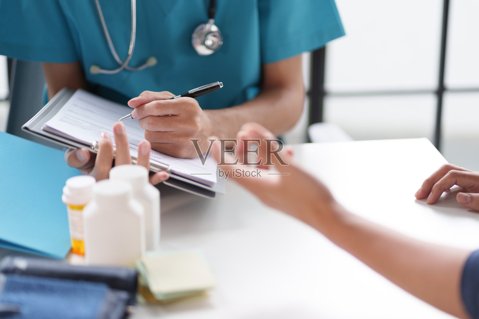 一位穿着蓝色制服的医生在与患者咨询时做笔记。桌子上有医疗瓶和文件。照片摄影图片