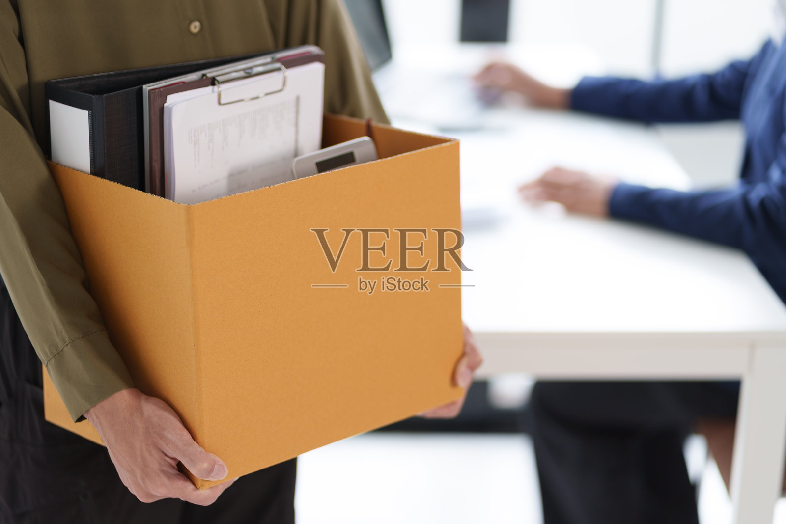 一个人提着装满办公用品的箱子，里面包括一个夹板和文件夹。这个场景暗示着搬家或过渡，可能与工作变动有关。照片摄影图片