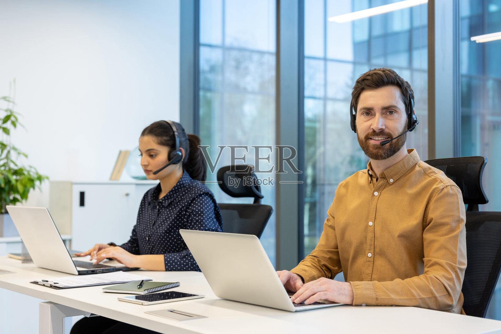 客户服务支持工作人员的肖像，戴着耳机的男士微笑着看向镜头，团队在办公室内使用笔记本电脑工作。照片摄影图片