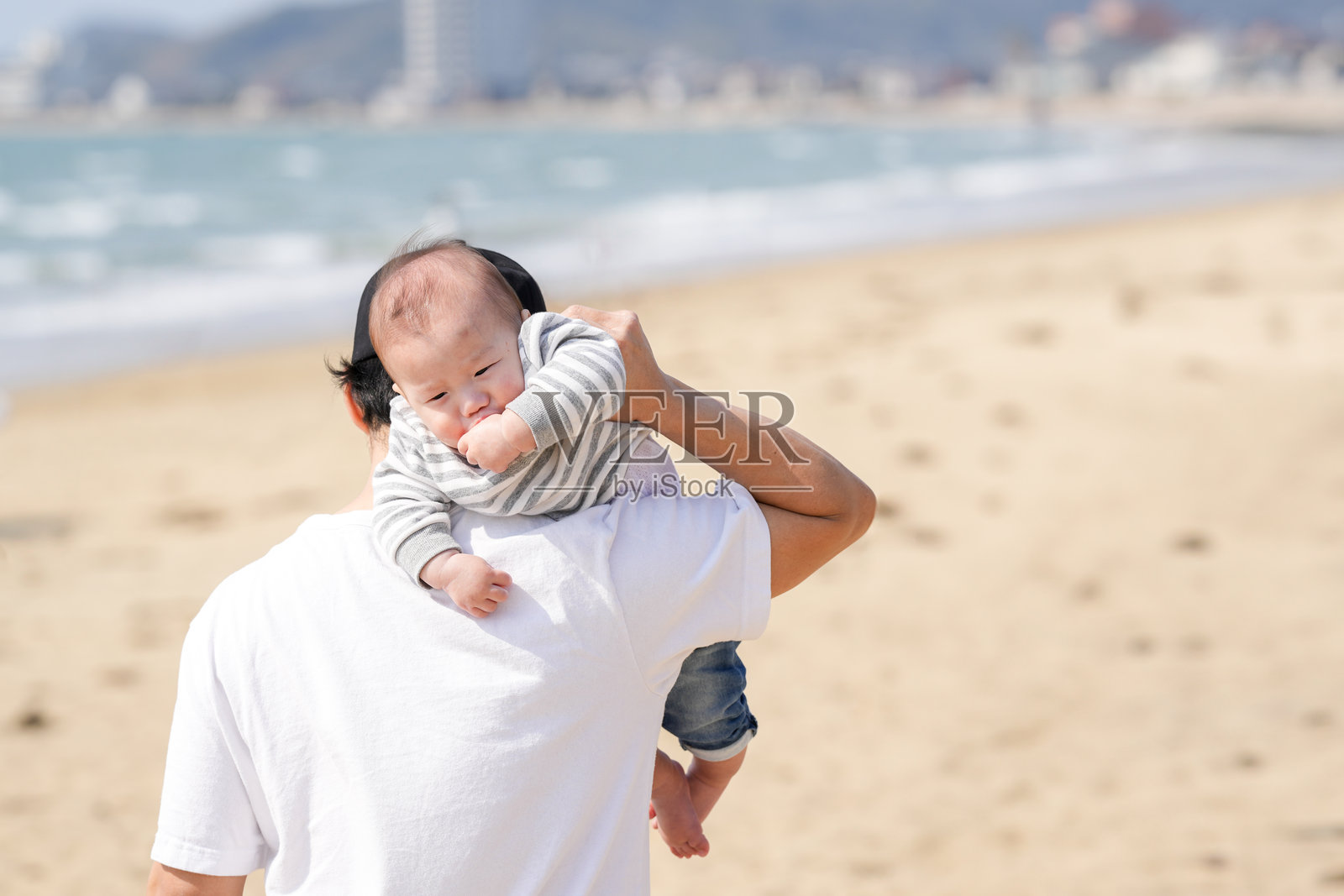 三月初，一位30多岁的日本父亲在海边的沙滩上背着他6个月大的宝宝，温暖的春季场景充满了家庭、快乐和亲密感。照片摄影图片
