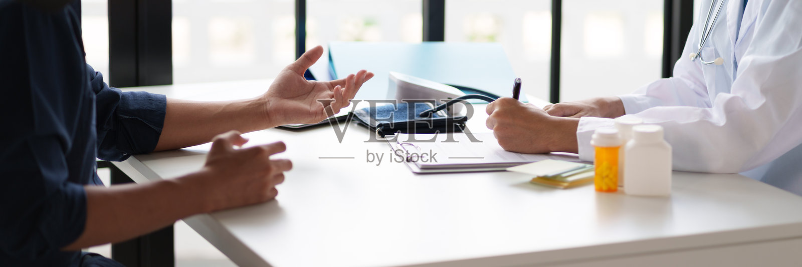 医生与患者在咨询期间。医生正在写字板上记录，而患者正在解释他们的症状。桌子上可见药瓶和血压监测仪。照片摄影图片